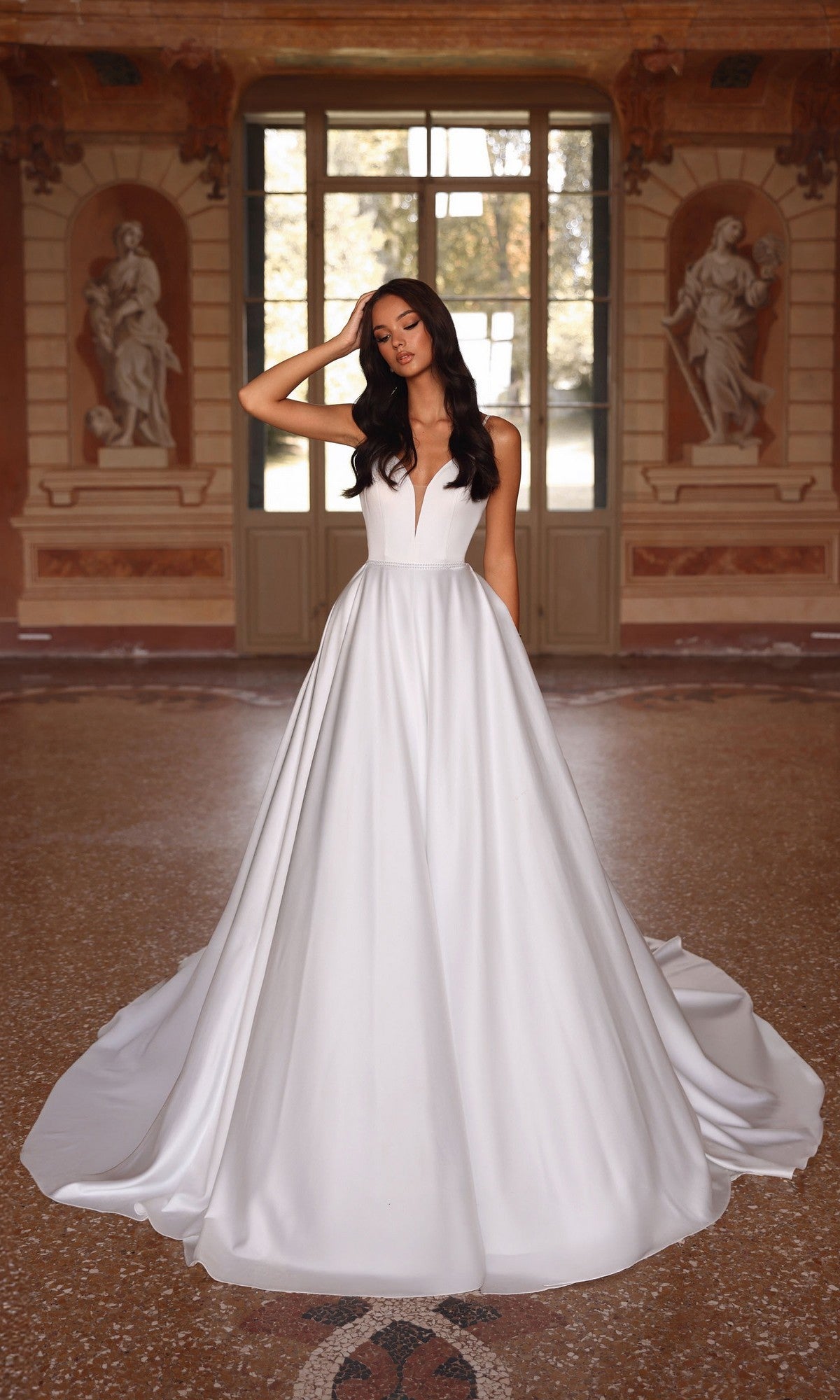 A woman wearing the Long Formal Dress: Alyce 7024 in diamond white, featuring a strapless bodice and elegant A-line skirt, stands in an ornate room with large windows and classical statues.